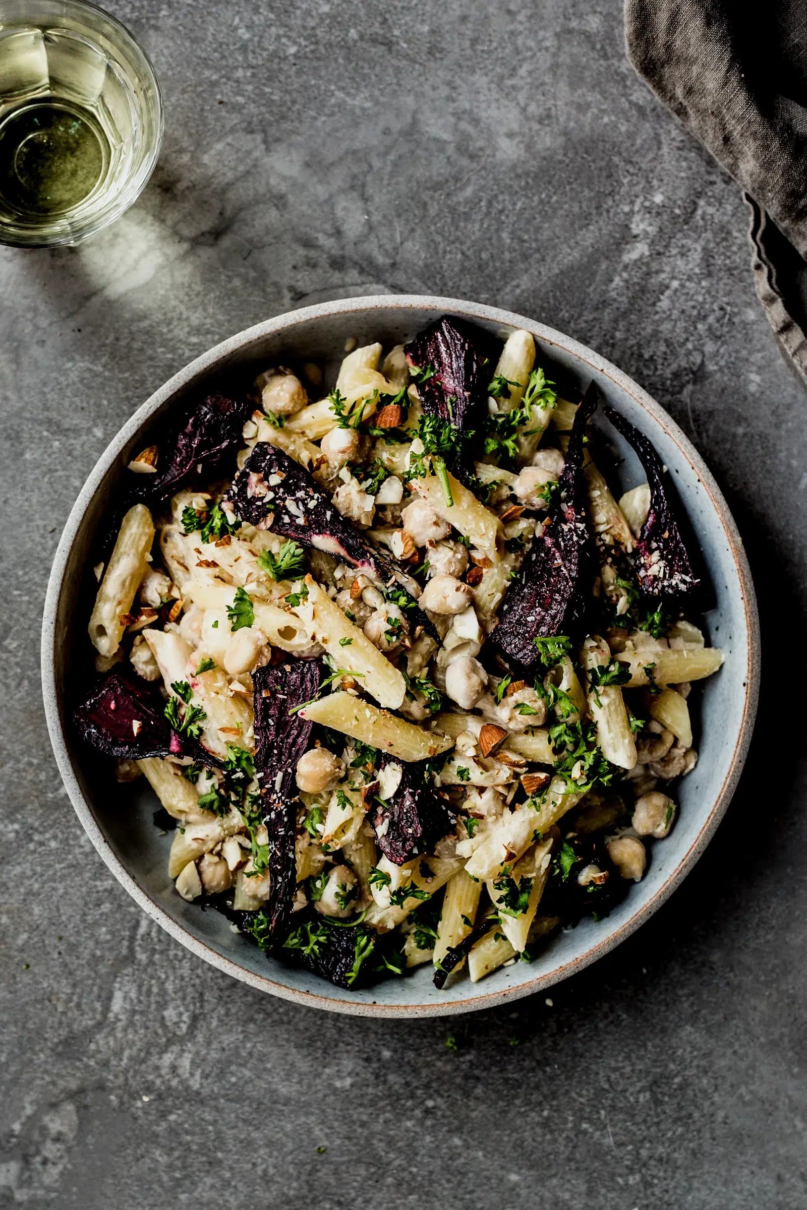 Bowl of chickpea pasta topped with roasted beetroot, fresh parsley, crushed almonds, and creamy sauce, served on a grey stone surface.