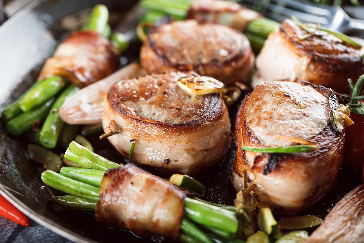 Pan-seared bacon medallions served with green beans wrapped in bacon, garnished with herbs and lemon, in a rustic skillet setting.