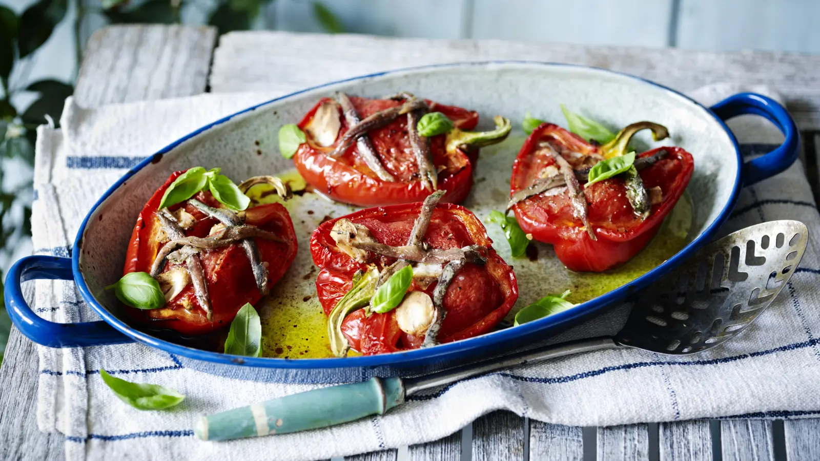 Stuffed romano peppers baked in a blue enamel tray, topped with anchovies, garlic, basil leaves, and olive oil, served on a rustic wooden table.