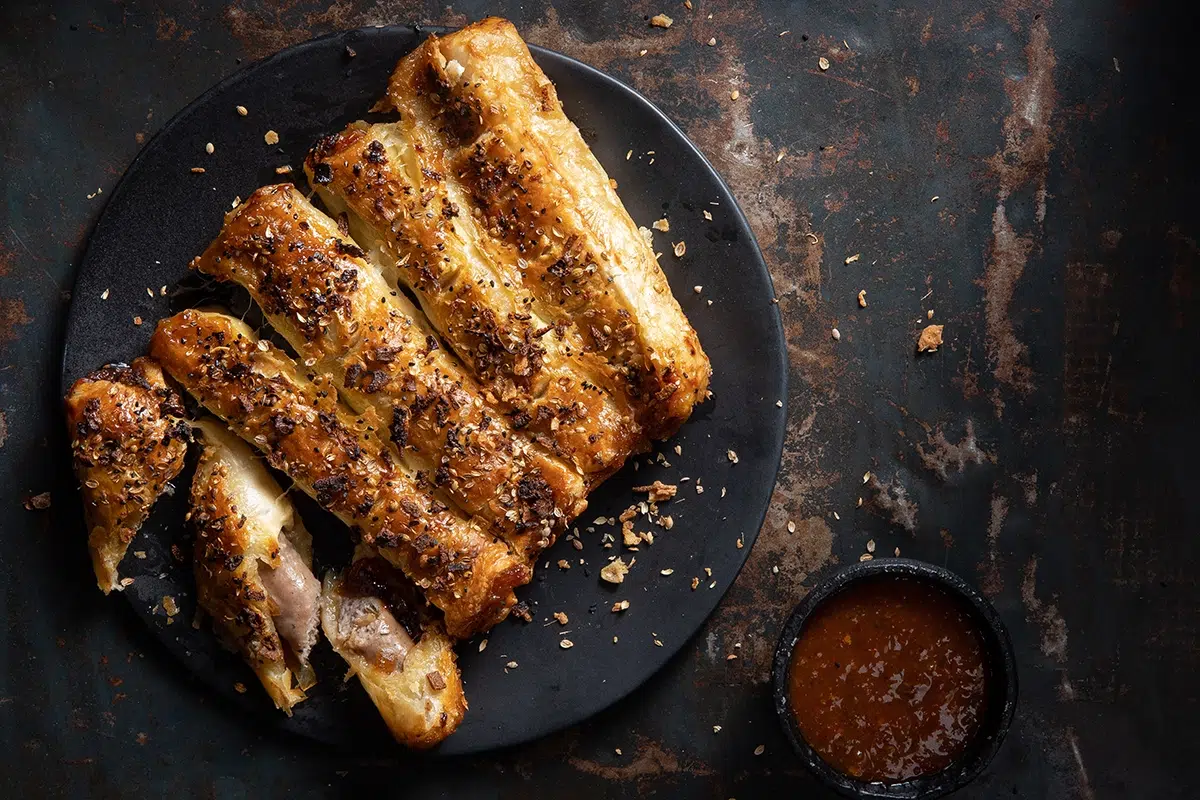 Golden air fryer sausage rolls with flaky pastry and seasoning, served on a black plate with a side of dipping sauce on a rustic background.