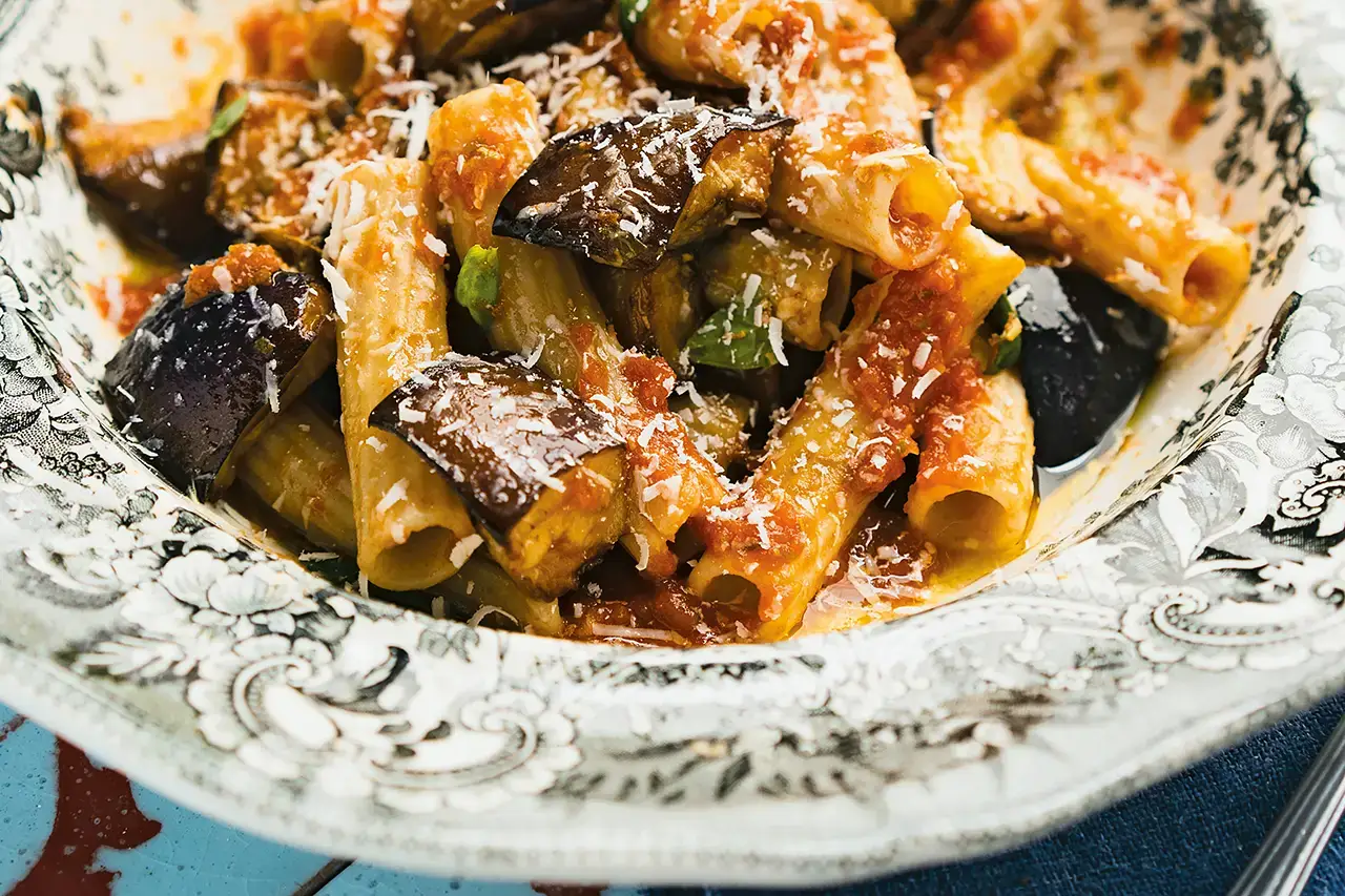 “Close-up of rigatoni pasta with roasted aubergine and tomato sauce, topped with grated cheese and fresh basil, served in a decorative porcelain bowl.”