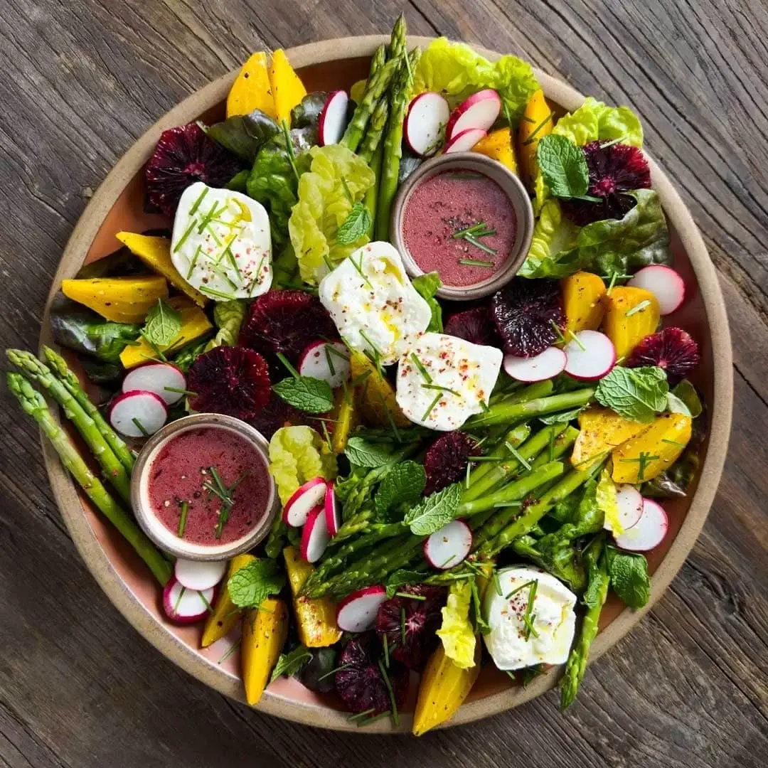A colourful beet salad featuring golden and red beets, asparagus, radishes, fresh mint, and small bowls of beet vinaigrette, served on a rustic wooden table without goat cheese.
