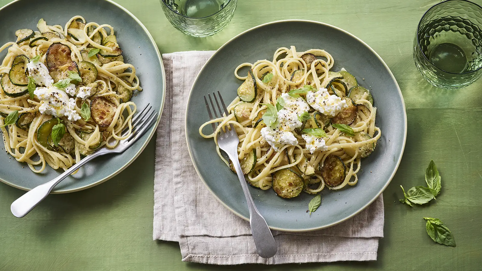 Two plates of linguine with sautรฉed courgette slices, fresh ricotta, black pepper, and basil leaves, served on a green wooden table with drinking glasses.