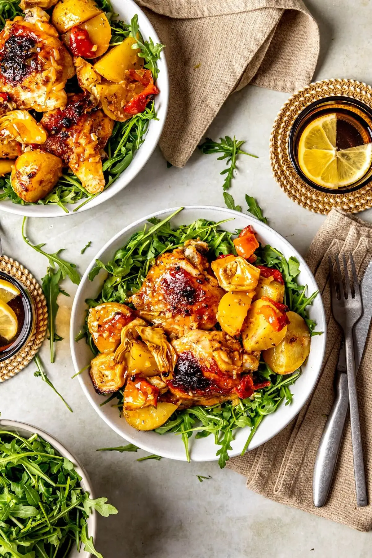 Overhead view of Greek lemon chicken with roasted potatoes, served on a bed of arugula in white bowls, accompanied by lemon water and napkins.
