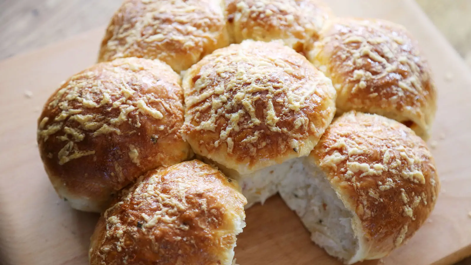 Close-up of freshly baked morning rolls with golden crusts and a dusting of grated cheese on top, arranged in a pull-apart cluster on a wooden board.