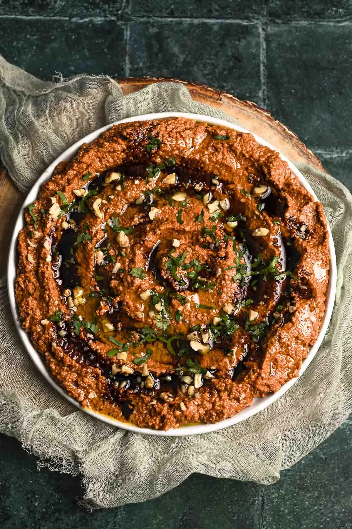 Overhead view of a bowl of muhammara dip garnished with chopped walnuts, parsley, and olive oil, served on a rustic green stone surface with linen cloth.