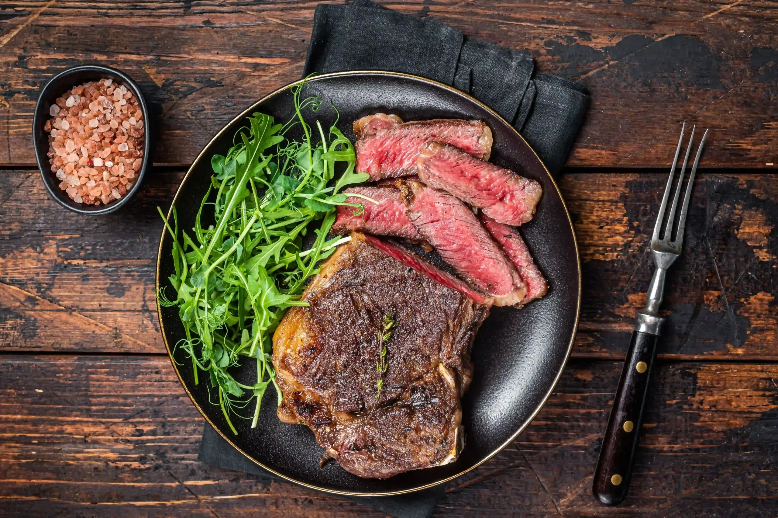 Sliced medium-rare sirloin steak served on a black plate with fresh rocket leaves, pink Himalayan salt on the side, and a vintage fork on rustic wooden table.