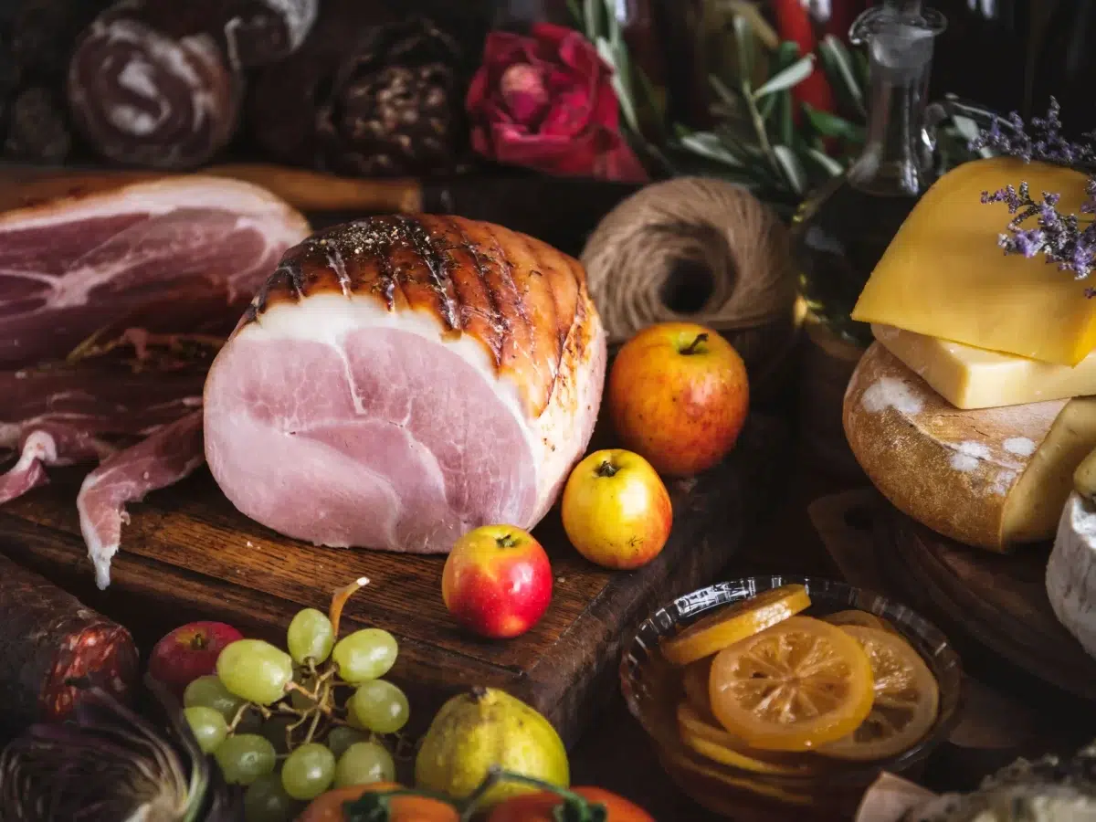 Rustic food scene featuring a glazed gammon joint surrounded by cheddar cheese, apples, grapes, preserved lemon slices, and cured meats — perfect for illustrating bold gammon steak pairings.