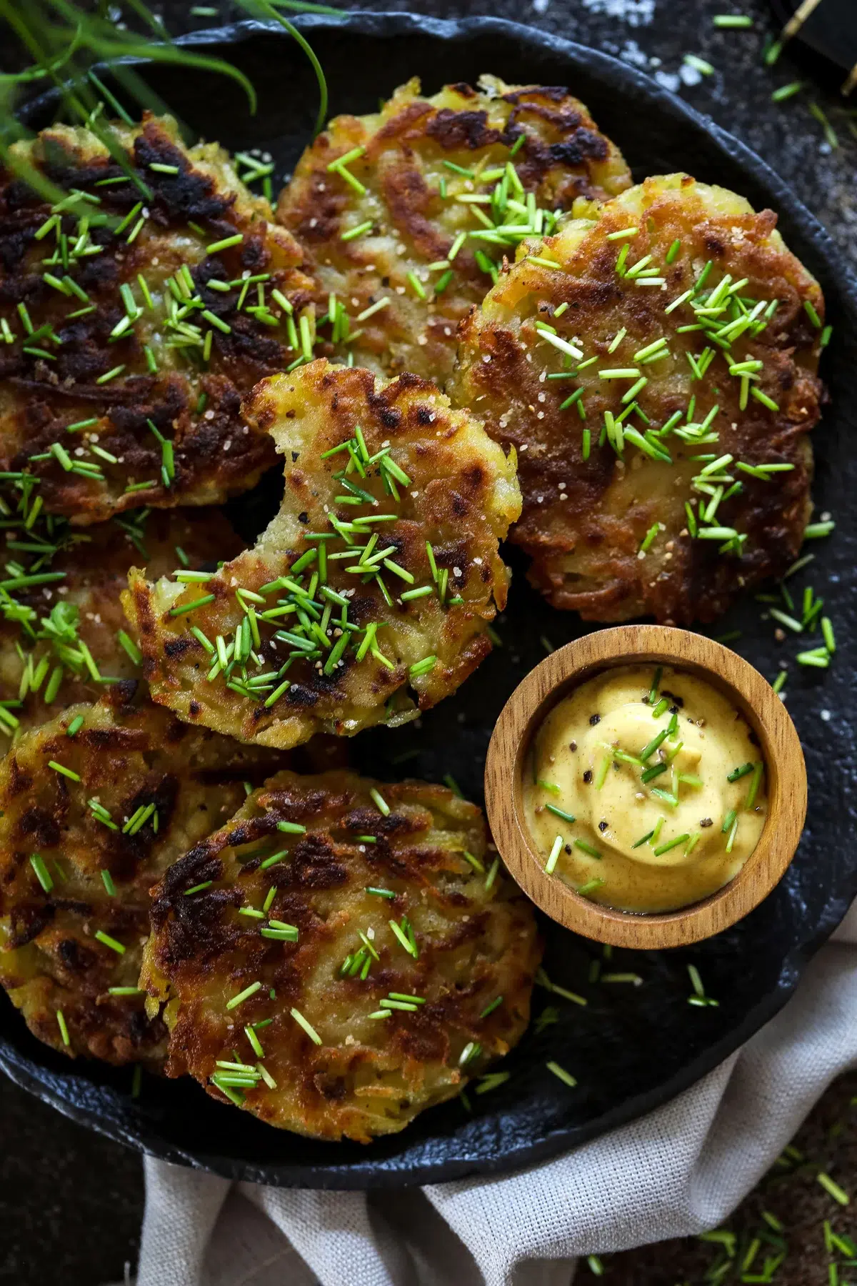 Close-up of crispy golden potato fritters garnished with fresh chives, served on a black plate with creamy dipping sauce in a wooden bowl.