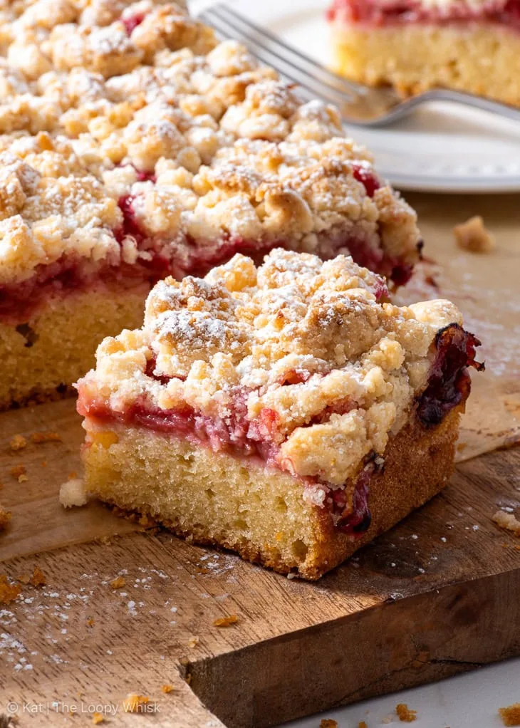 Close-up of a moist and crumbly slice of old-fashioned rhubarb cake topped with golden streusel and dusted with powdered sugar, served on a rustic wooden board.