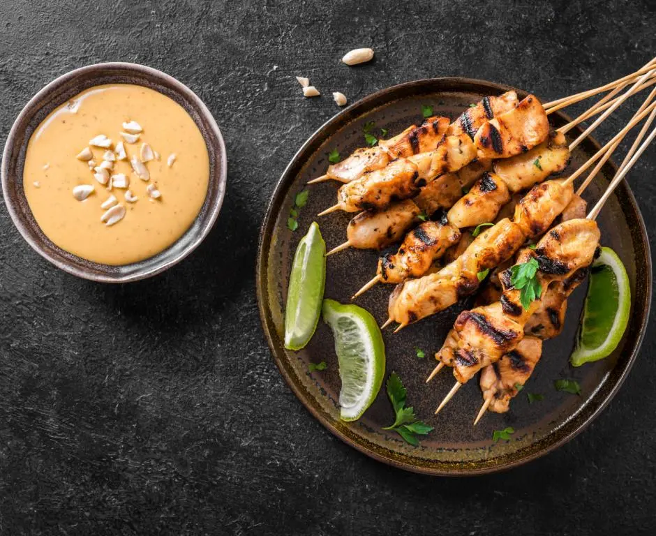 Grilled Thai chicken satay skewers served with creamy peanut sauce and lime wedges on a dark rustic background.