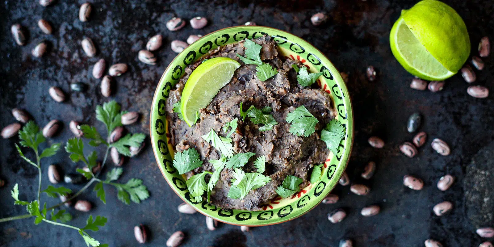Bowl of creamy refried refried beans garnished with fresh cilantro and lime, surrounded by pinto beans on a rustic dark background.