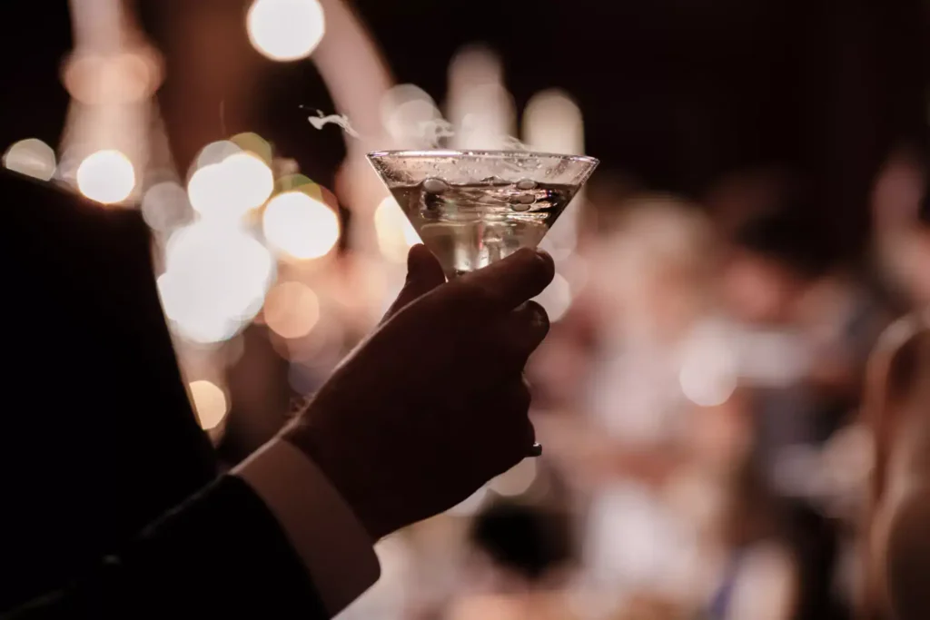 Elegant hand holding a chilled vodka martini glass at a glamorous evening event, with blurred golden lights in the background.
