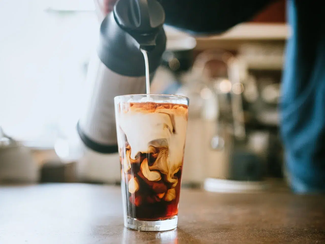 Close-up of a TikTok-inspired iced latte with milk being poured into cold coffee, creating a beautiful swirl effect in a clear glass on a café counter.