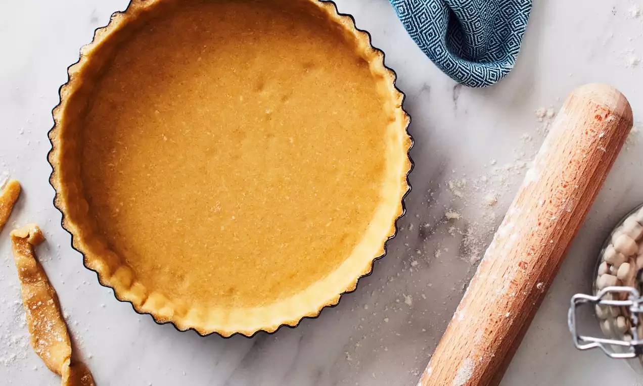 A perfectly blind-baked sweet pastry shell in a fluted tart tin, with a wooden rolling pin and baking beans visible nearby—ready for filling with sweet tarts or pies. Read our sweet pastry recipe