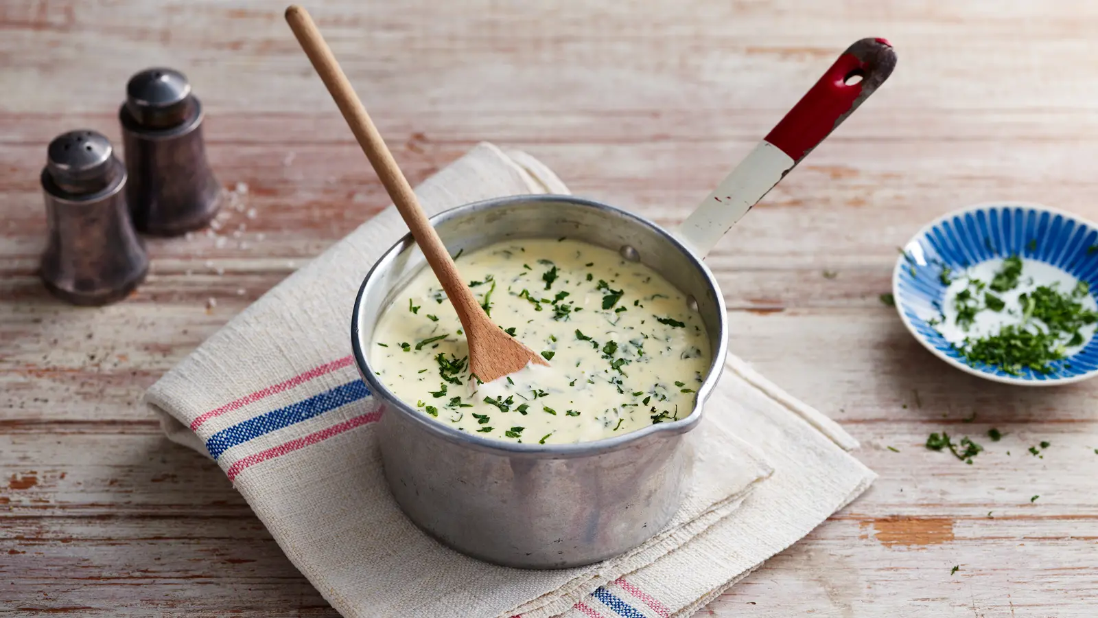 "Creamy parsley sauce in a small metal saucepan with a wooden spoon, topped with freshly chopped parsley, placed on a folded kitchen towel on a rustic wooden table."