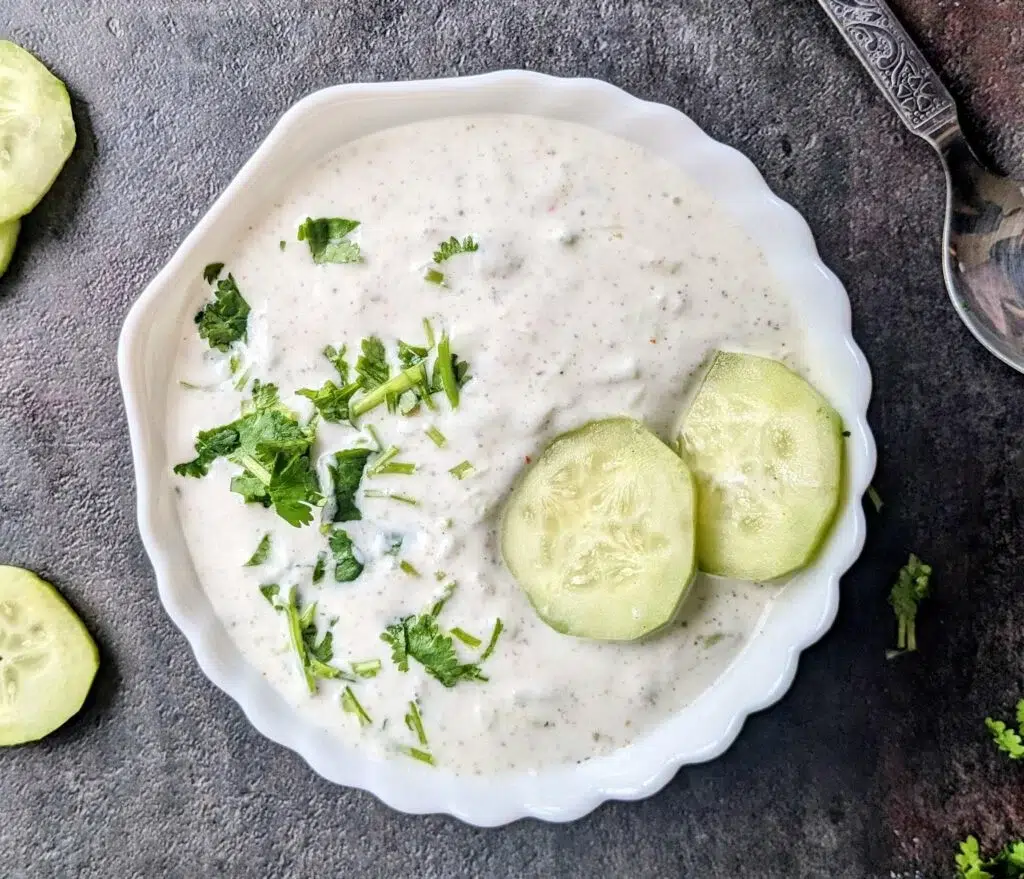 Cucumber Raita Recipe served in a white bowl, topped with fresh coriander leaves and cucumber slices, on a dark stone surface.