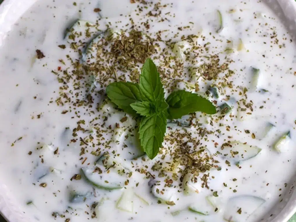 "Cucumber Raita Recipe in a white bowl, garnished with fresh mint leaves and sprinkled with dried herbs, ready to serve with Indian dishes."
