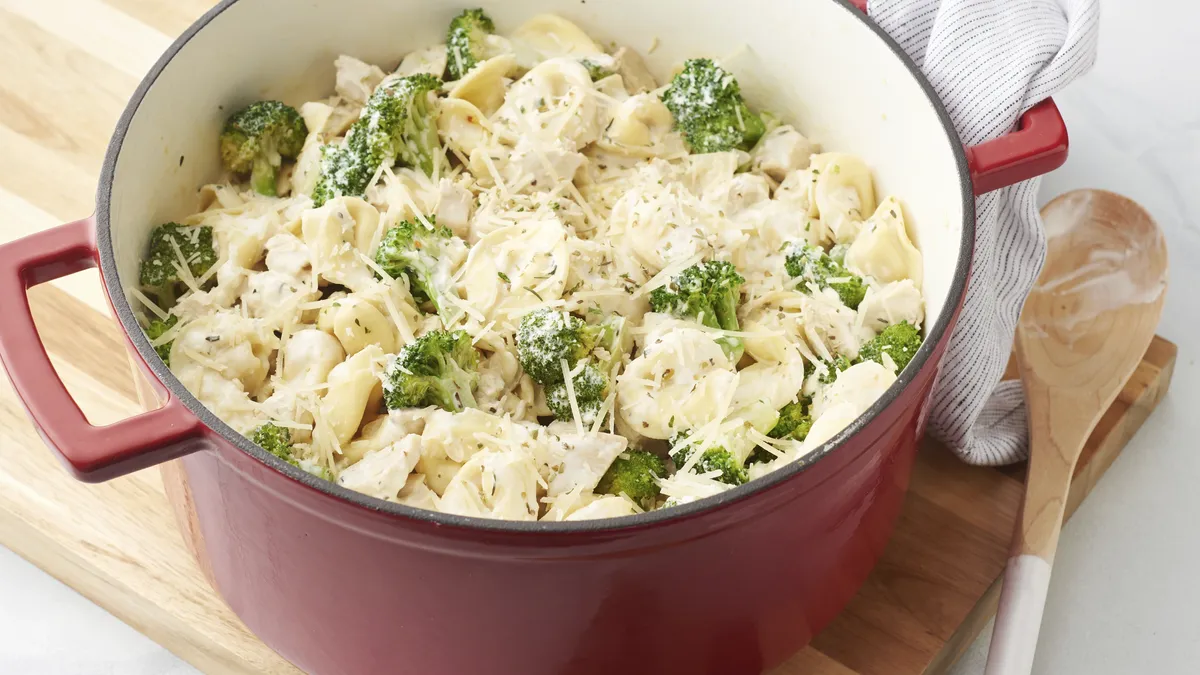 Red cast-iron pot filled with creamy cheese pasta, broccoli florets, and shredded Parmesan on a wooden board with a wooden spoon and striped cloth next to it.