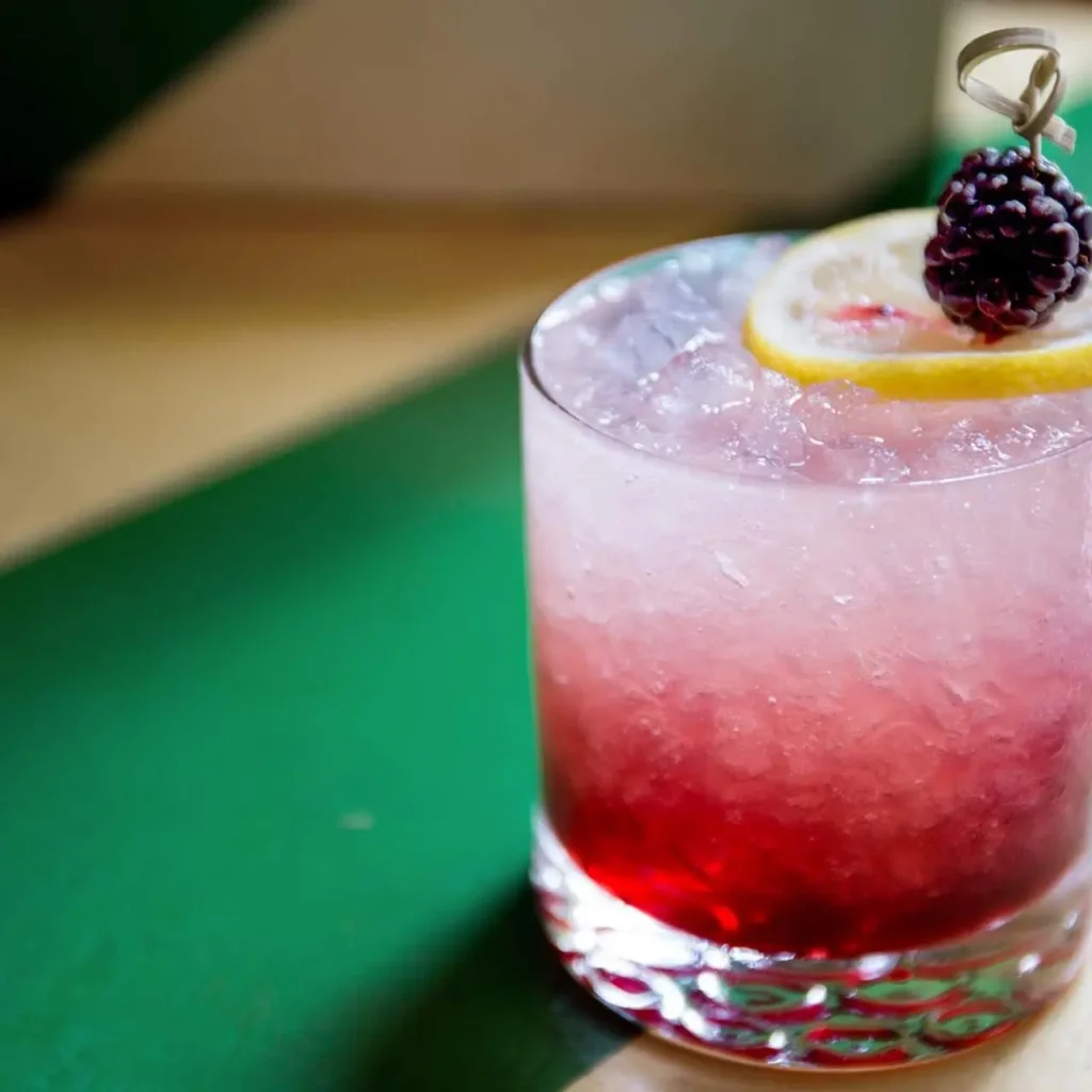 Bramble cocktail served in a short glass with crushed ice, featuring a gradient from pink to deep purple, garnished with a lemon slice and a fresh blackberry on a cocktail pick.