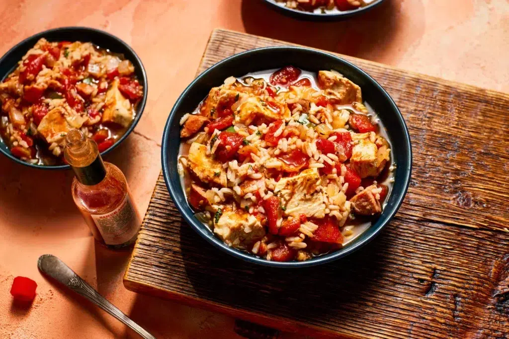Close-up of a hearty slow cooker chicken chorizo jambalaya served in a dark bowl, featuring tender chicken chunks, spicy chorizo slices, fluffy rice, and diced tomatoes, with a bottle of hot sauce on the side.