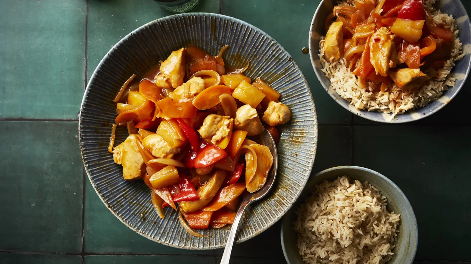 Authentic sweet and sour Hong Kong style chicken served in a blue bowl with chunks of pineapple, red and orange bell peppers, and onions in a glossy sauce, accompanied by bowls of steamed rice.