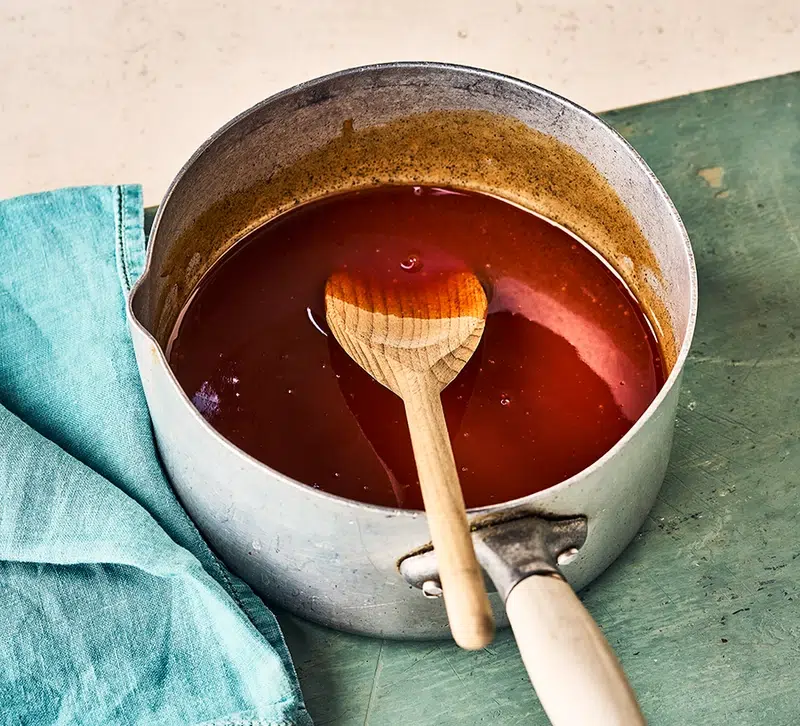 Glossy sweet and sour sauce simmering in a saucepan with a wooden spoon resting inside, ready to serve.