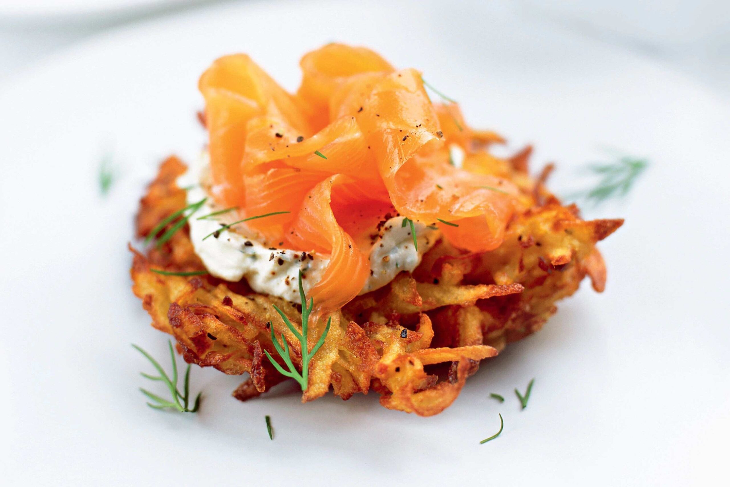 Crispy potato rösti topped with creamy dill sauce and thin slices of smoked salmon on a white plate.